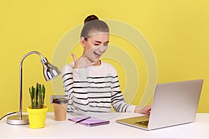 Woman sitting at workplace, keeping hand shaped like telephone near head, looking at laptop screen.