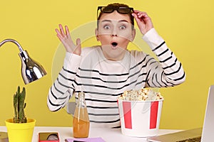 Woman sitting at workplace with popcorn and beverage, raising glasses and watching shocking moment.