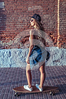 Woman on skateboard, summer in city, brick wall background. Denim baseball cap long hair. The concept of fashion style