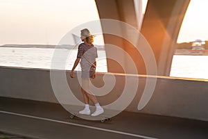 Woman skateboarder on longboard on street at sunset. Female longboarder riding alone alongside river