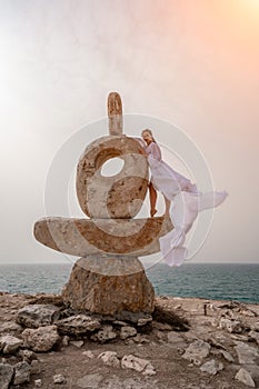 Woman sky stone. Lady stands on stone sculpture with ocean view. She is dressed in a white long dress, against the