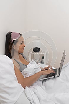 woman in sleep mask works from home in bed, using her laptop to surf internet and study remotely.