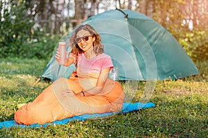 Woman in sleeping bag drinks pure water against the backdrop of tent in a camping site at early morning