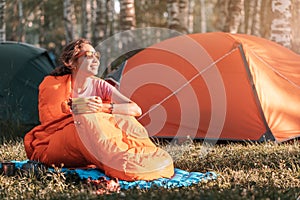 Woman in sleeping bag eating breakfast against the backdrop of many tents in a camping site at early morning