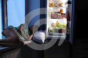 Woman sleeping on window sill near open refrigerator in kitchen