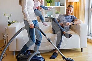 Woman with small child doing housekeeping while man sitting in couch