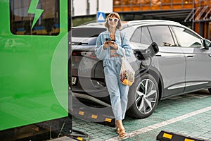 Woman with a smart phone and groceries waiting for her electric car to be charged outdoors
