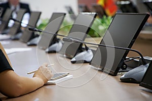 A woman with a smartphone, fountain pen and notebook sits at a table with monitors and intercoms during a conference call. Without