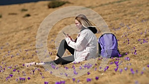 Woman with smartphone sitting on valley of high mountain among crocus flower. Lady traveler with backpack using mobile