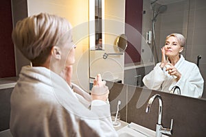 Woman smearing face with cream and looking at herself in mirror in bathroom