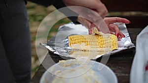 A woman smears corn with oil and spices to bake in foil over a fire. Hands and corn close-up. Motion shooting on the table