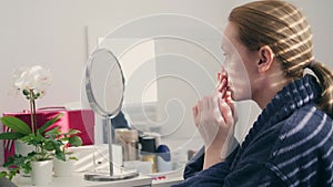 a woman smears her face with cream at home sitting in front of a mirror. a shadow falls on her from the blinds