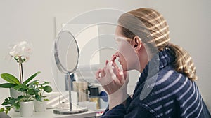 a woman smears her face with cream at home sitting in front of a mirror. a shadow falls on her from the blinds