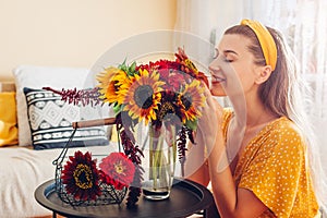 Woman smells bouquet of sunflowers with zinnia flowers arranging in vase at home. Lady enjoys fresh blooms. Interior