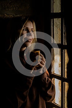 A woman smiles and drinks coffee by the window in the dark. A woman in her forties