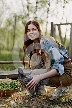 Woman smiles looking at the chicken she holds near the feeder in her hands on the farm, farm labor for raising healthy