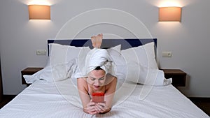 Woman smiling browsing in smartphone lying in bed with towel on hair after bath.