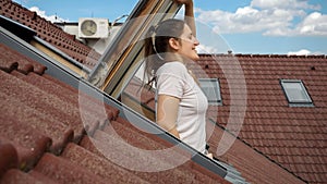 Woman smiling while looking out of an attic window under a red tiled roof and breathing fresh air