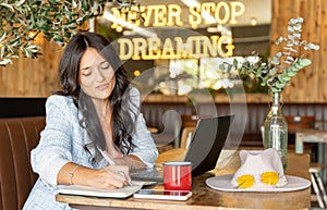 Woman smiling and writing on a notebook while using a laptop in a coffee shop