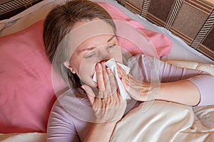 Woman sneezes, has a running nose or seasonal allergy. Middle aged 40 years woman lying in the bed and holds napkin tissue near