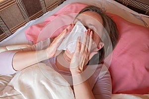 Woman sneezes, has a running nose or seasonal allergy. Middle aged 40 years woman lying in the bed and holds napkin tissue near