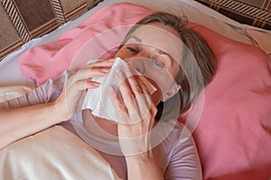 Woman sneezes, has a running nose or seasonal allergy. Middle aged 40 years woman lying in the bed and holds napkin tissue near