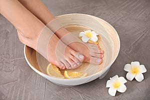 Woman soaking her feet in bowl with water, orange slices and flower on grey background, closeup.