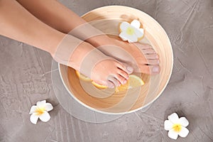 Woman soaking her feet in bowl with water, orange slices and flower on grey background, top view