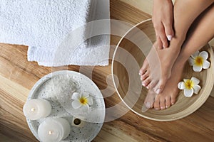 Woman soaking her feet in dish with water and flowers on wooden floor, top view.