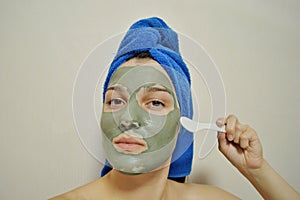 Woman with a spatula applies a clay mask to her face with blue towel on her head