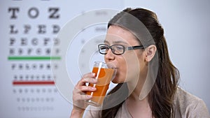 Woman in spectacles drinking fresh carrot juice, eyechart on background, vision