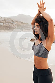 Woman in sports clothes standing with hands joined overhead. Female meditating at the beach with closed eyes