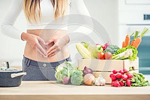 Woman with sports figure on her belly shows heart shape in home kitchen with wooden box full of organic vegetable