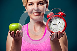 Woman in sportswear holding alarm clock and apple. People, fitness and healthcare concept