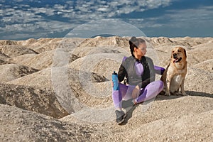 Woman in sportswear sits on yellow dunes with dog labrador retriever looks each other on sunny day, copy space, concept