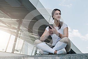 Woman in sportswear sitting in city street, relax after sports training, use smartphone, listen to music, checking email
