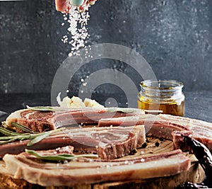 A woman sprinkles salt on a Raw Lamb Ribs with the ingredients for cooking: pepper, olive oil and herbs on a wooden stand