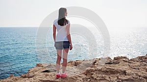 Woman stand at edge of cliff on sunny day in slow motion. Carefree female look at view of blue sea