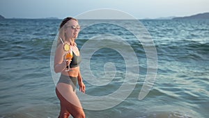 Woman stand knee-deep in sea on sunset. Girl on beach in green swimsuit having fun, drinks her orange cocktail, big