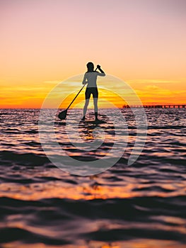 Woman stand up paddle boarding at dusk on a flat warm quiet sea with beautiful sunset colors