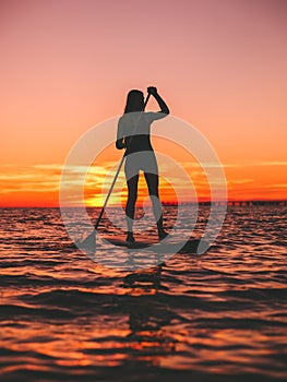 Woman stand up paddle boarding at dusk on a flat warm quiet sea with beautiful sunset