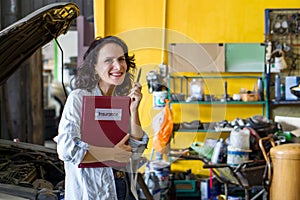 A woman standing in an auto repair shop holding a red folder with the word Insurance written on the cover.