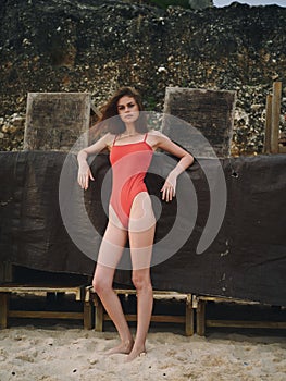 Woman standing on the beach on the sand in a red bathing suit near the rocks, beautiful slim body with a tan