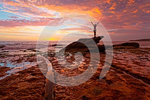 Woman standing on coastal rocks with stunning sunrise and reflections