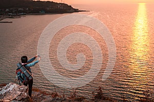 A woman is standing on the edge of a cliff and taking photos of the sunset on her smartphone. In the background, the sea at sunset