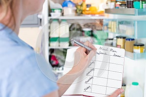 Woman Standing In Front Of Refrigerator In Kitchen With Notebook Writing Weekly Meal Plan