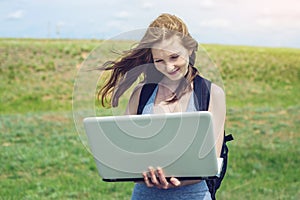 Woman standing on a green field on the background of sky with clouds and working or studying with laptop wireless