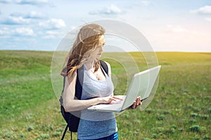 Woman standing on a green field on the background of sky with clouds and working or studying with laptop wireless