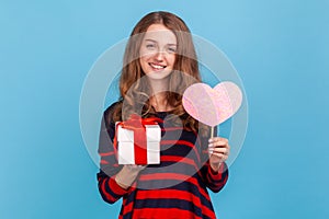 Woman standing holding present box and showing pink heart on stick, looking at camera with love.