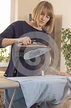 Woman is standing at an ironing board and holding an iron from which steam escapes. Woman during household chores. Ironed clothes